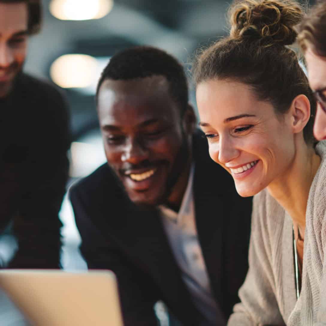 Group of diverse professionals smiling and collaborating around a laptop in a modern office.