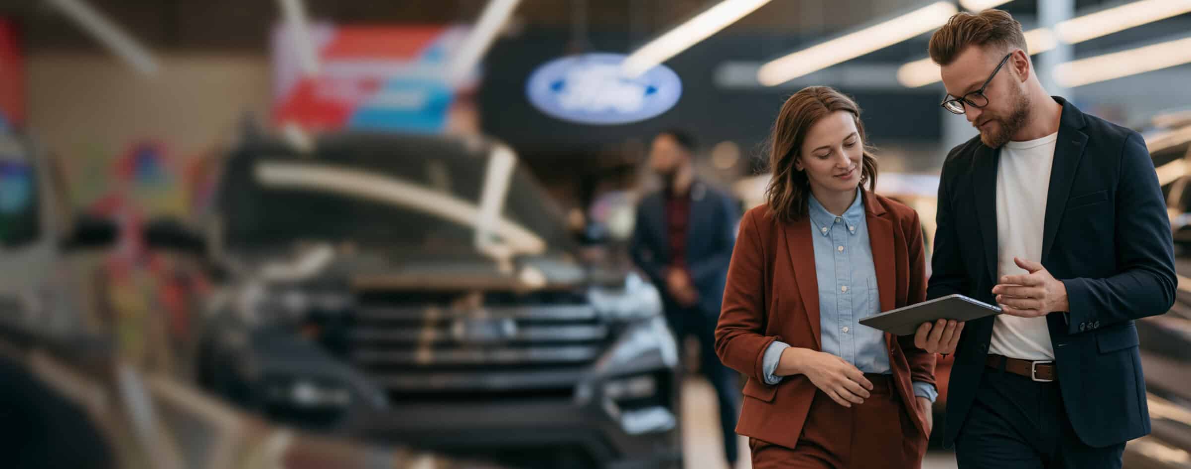 Two professionals discussing data on a tablet in a car dealership showroom.