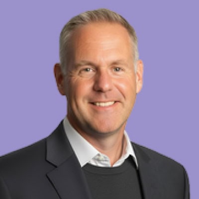 Smiling middle-aged man in a suit with a purple background, looking directly at the camera.
