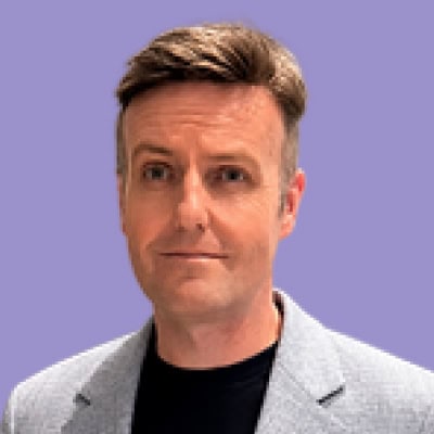 Professional portrait of a man with short brown hair, wearing a gray blazer and black shirt, against.